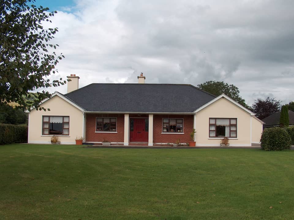 A single-storey rural bungalow with two cream-rendered front gables, a red-brick recessed porch with a red door, a slate roof and a large lawn in front, under a cloudy sky.