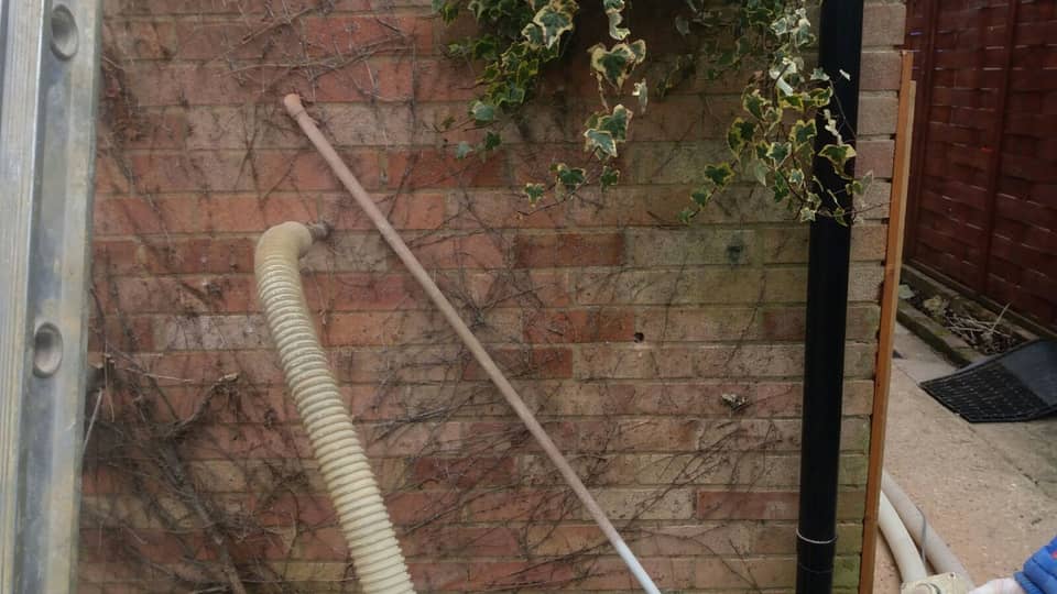 A ribbed insulation hose plugged into a drilled hole in an ivy-covered red-brick house wall, with a second drilled hole visible to the right and a ladder at the edge of the frame.