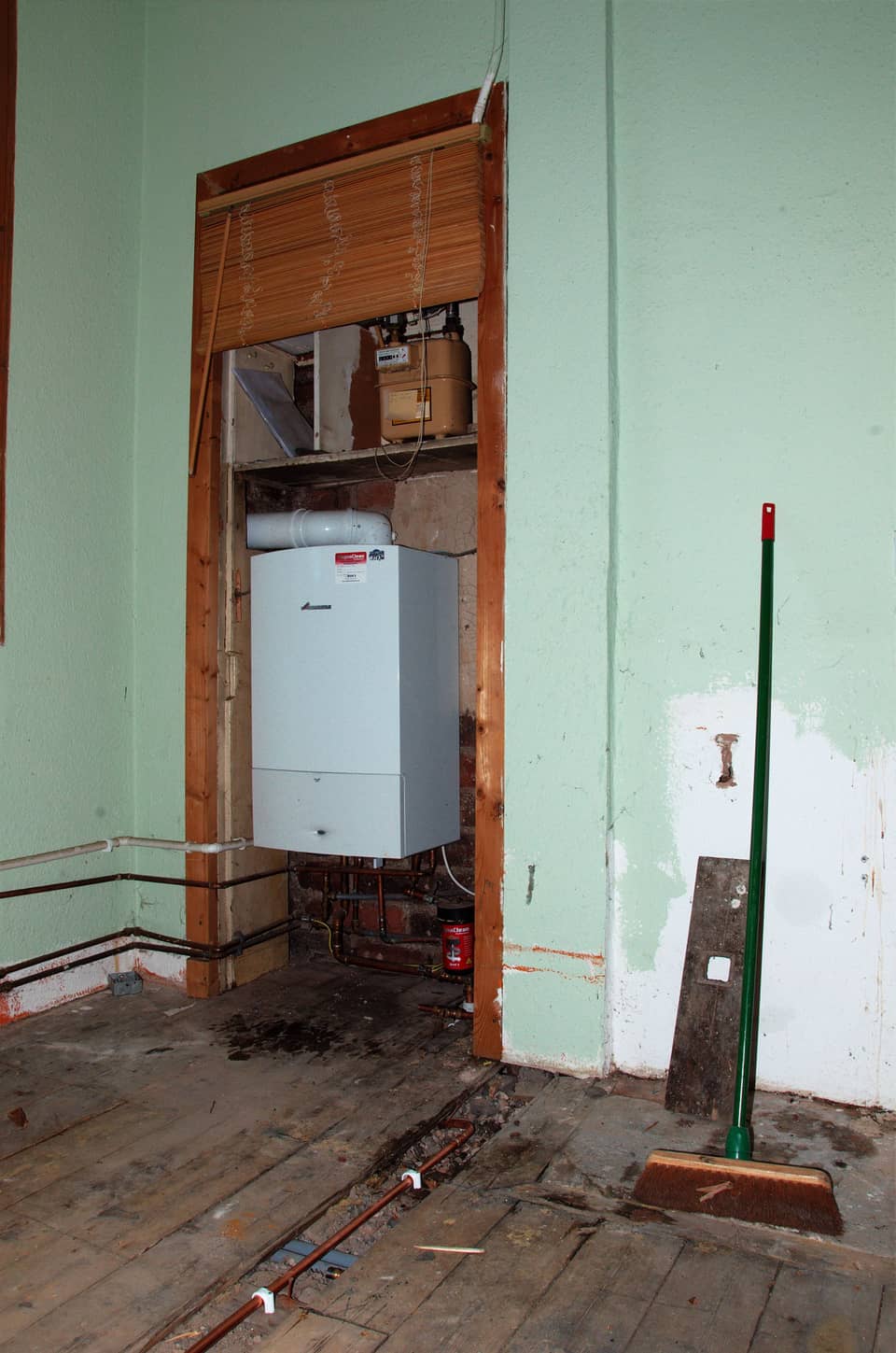 A new white Worcester wall-hung gas boiler fitted in an alcove beside the gas meter, in a room stripped back for a kitchen refit with floorboards lifted and copper pipes exposed.