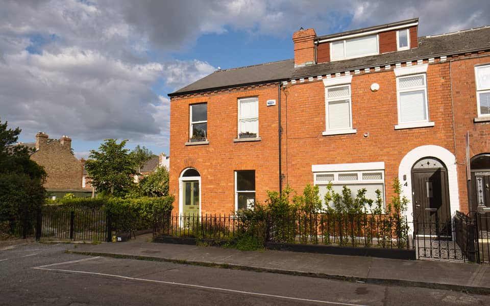 An end-of-terrace Victorian red-brick house with arched doorways, iron railings and a later attic dormer, on a quiet Drumcondra street in evening sun.