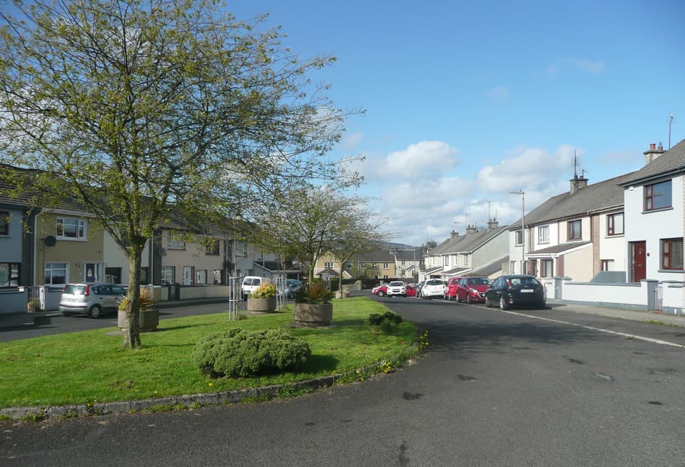 A housing estate street in Letterkenny: dry-dash semi-detached houses on both sides, cars at the kerb, a grass island with a young tree, hills at the end of the road.