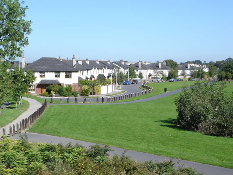 A curving road of white-rendered two-storey semi-detached and terraced houses with dark tiled roofs, set behind a large green and a footpath, under a clear blue sky.