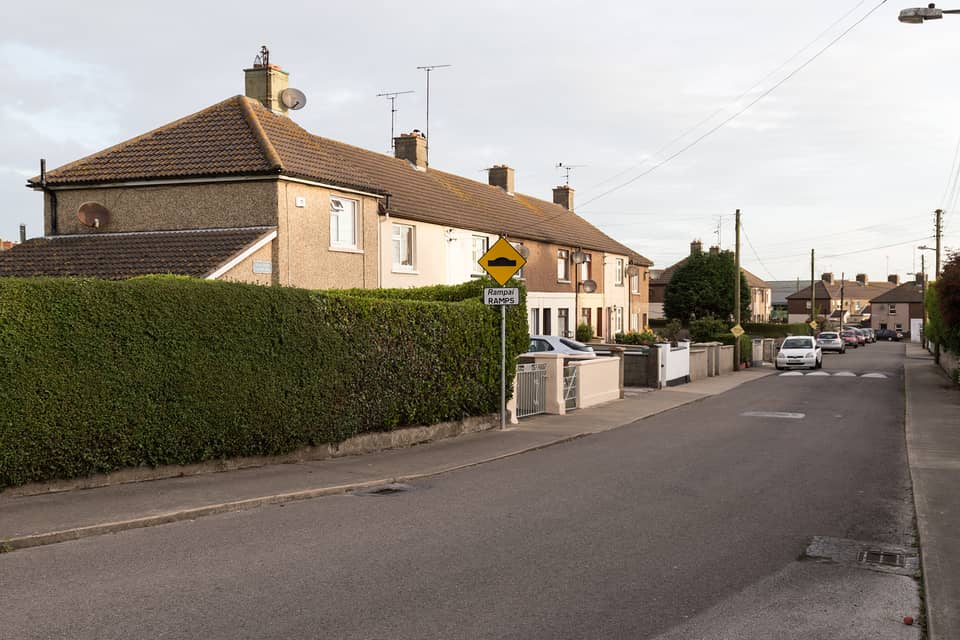 A row of two-storey terraced houses with hipped tiled roofs and a mix of pebble-dash and smooth render behind a tall hedge on Dolphin Road, with a bilingual “Rampaí / Ramps” sign.